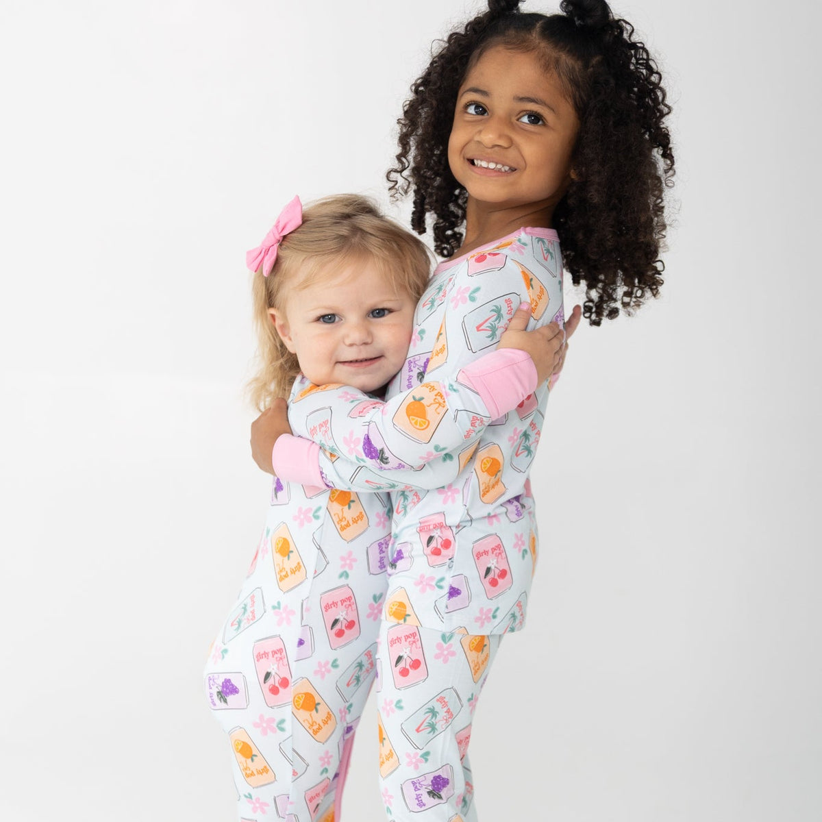 Two young children hugging while wearing matching long-sleeve pajamas with a light blue background with a colorful cherry, orange and grape soda cans print. set against a plain white studio background.