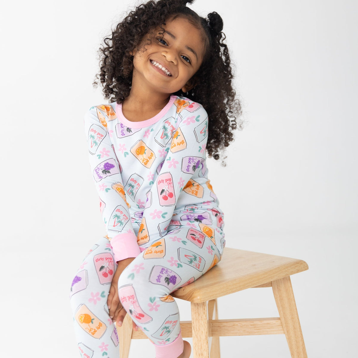 A young girl with curly black hair sits on a light wood stool wearing a light blue pajama set with a colorful cherry, orange and grape soda cans print. The background is a clean white studio backdrop.