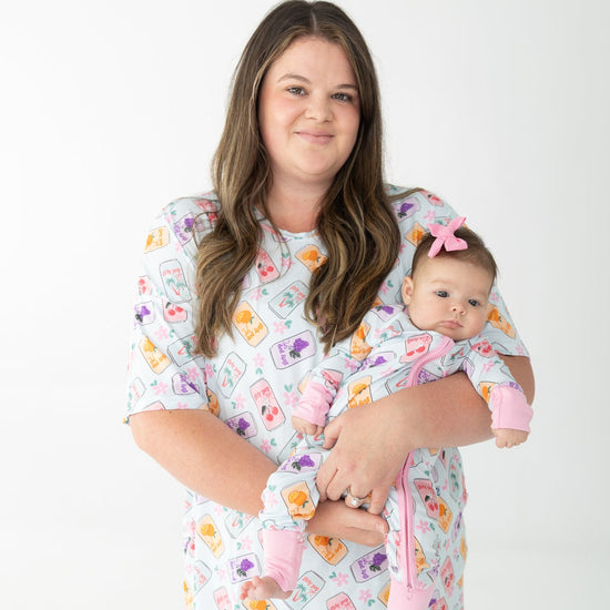 A woman holding a baby wearing matching printed pajamas, colorful repeating pattern of fruit and flowers, colorful soda cans pattern on a light blue background, photographed against a seamless white studio backdrop.