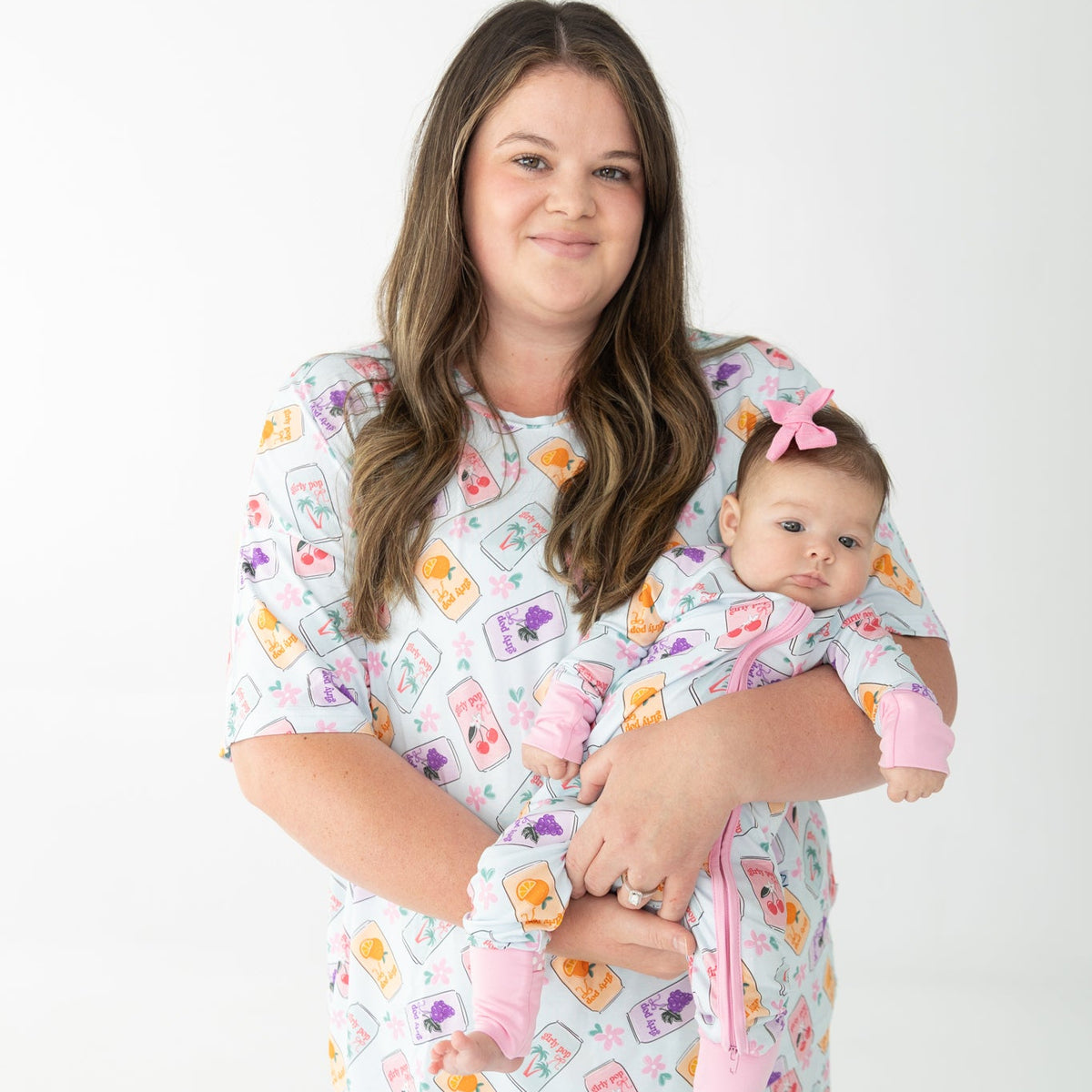 A woman holding a baby wearing matching printed pajamas, colorful repeating pattern of fruit and flowers, colorful soda cans pattern on a light blue background, photographed against a seamless white studio backdrop.