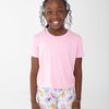 A young girl with curly black hair smiling, wearing a light pink short-sleeve t-shirt and light blue shorts with a colorful floral and soda can pattern.