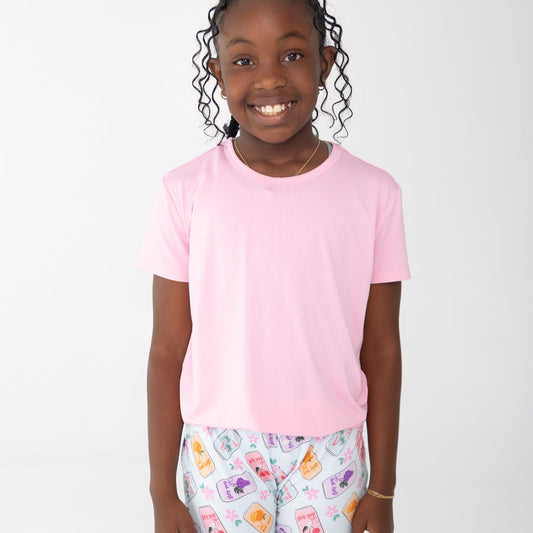 A young girl with curly black hair smiling, wearing a light pink short-sleeve t-shirt and light blue shorts with a colorful floral and soda can pattern.
