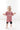The image shows a young toddler walking indoors on a light-colored wooden floor. The child is smiling brightly and wearing a mauve-pink oversized T-shirt with black shorts underneath. The T-shirt has white lettering across the front. The toddler has a white bow in their hair and appears to be mid-step, looking joyful and energetic. The background is minimal and softly lit, creating a clean and airy atmosphere.
