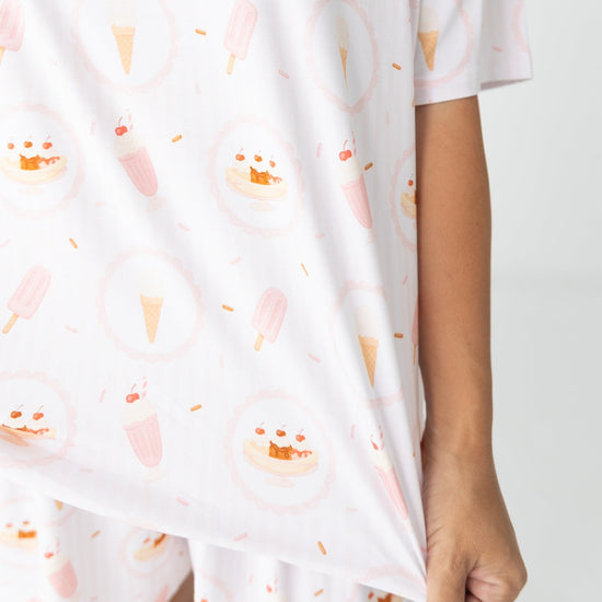 A close-up of a person wearing a white short-sleeve pajama top with a repeating pastel print of ice cream cones, popsicles, and ice cream sundae's paired with matching white pajama bottoms.