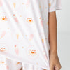 A close-up of a person wearing a white short-sleeve pajama top with a repeating pastel print of ice cream cones, popsicles, and ice cream sundae's paired with matching white pajama bottoms.