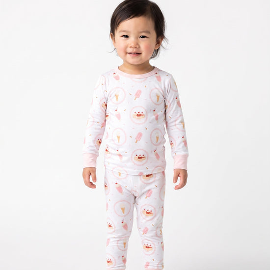 A toddler standing against a plain light gray studio background, wearing a long-sleeve pajama set with a white base and a repeating pink ice cream theme.