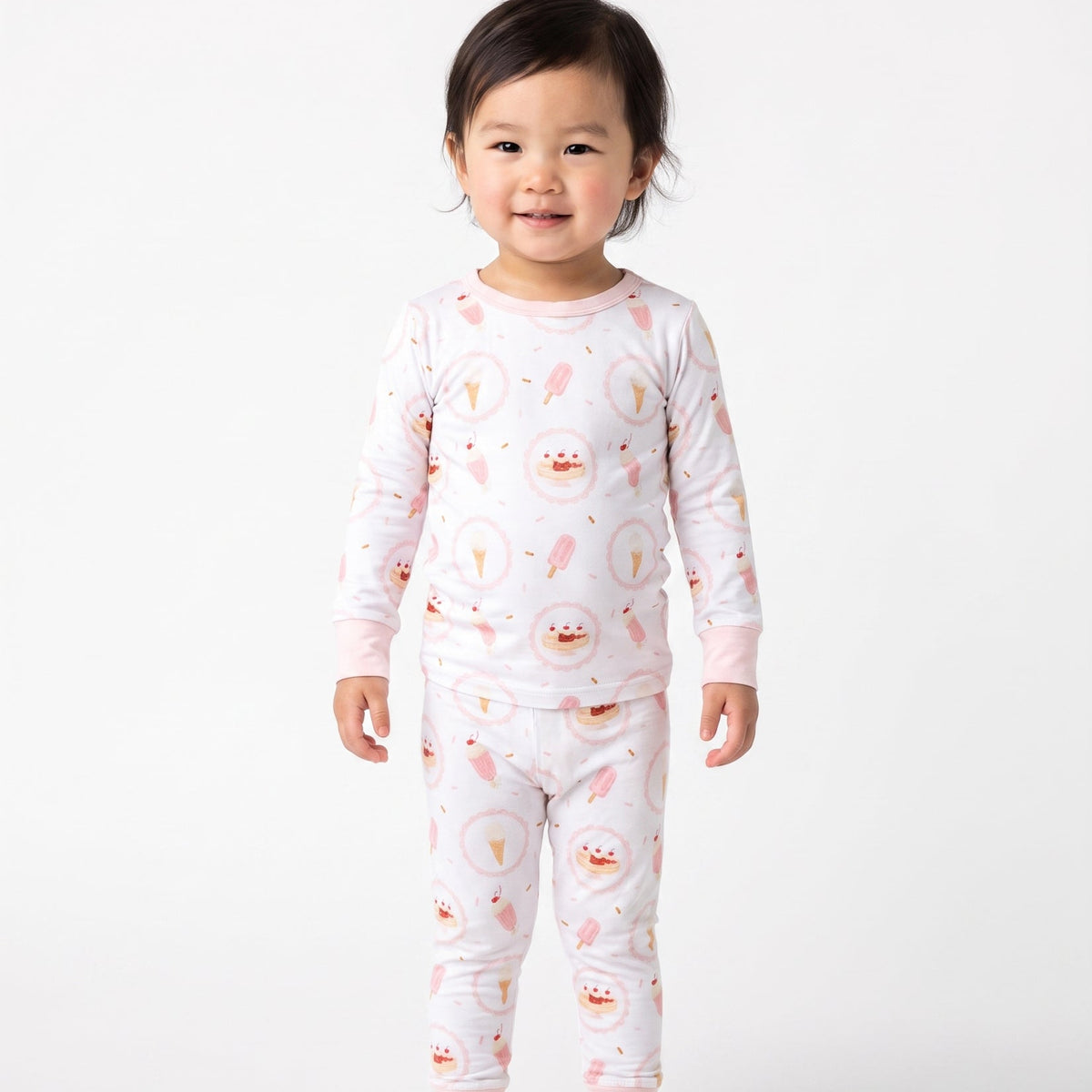 A toddler standing against a plain light gray studio background, wearing a long-sleeve pajama set with a white base and a repeating pink ice cream theme.