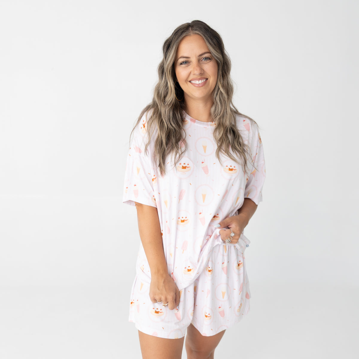 A woman modeling a matching short-sleeve t-shirt and shorts pajama set with a white base with pink accents, ice cream sundae, ice cream cone, ice cream bar, milkshake. 