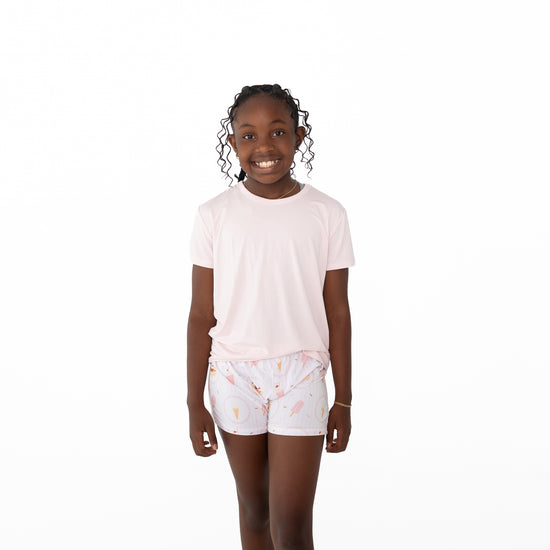 A young girl smiling and standing against a solid white background, wearing a light pink short-sleeve t-shirt and white shorts with a pastel pink ice cream print.