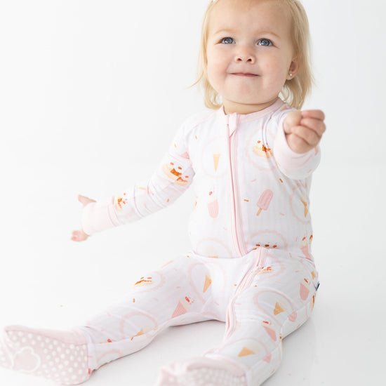 A baby girl sitting on a white studio background wearing a light pink footed zip-up sleeper with a pink ice cream bar, ice cream sundae, ice cream cone pattern.