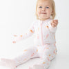 A baby girl sitting on a white studio background wearing a light pink footed zip-up sleeper with a pink ice cream bar, ice cream sundae, ice cream cone pattern.