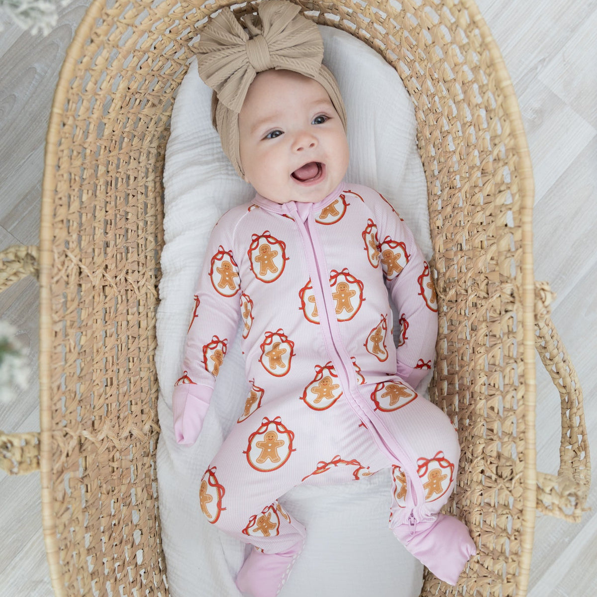 A baby lying in a woven straw Moses basket, wearing a pink and white striped gingerbread repeating print.