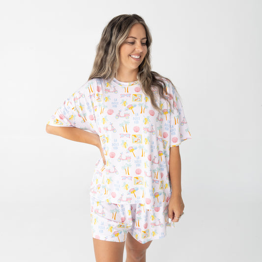 A woman with long blonde hair wearing a short-sleeve pajama set with a colorful tropical travel print on a white background, photographed against a solid white studio background.