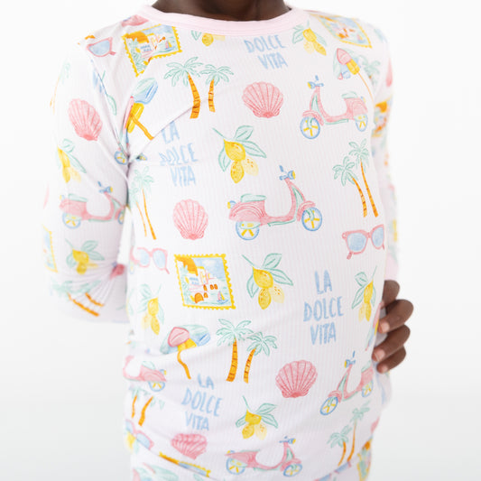 A child wearing a white long-sleeve top with a colorful 'La Dolce Vita' pattern featuring palm trees, pink scooters, seashells, lemons, and sunglasses.