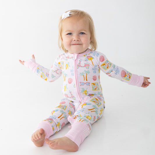 A baby girl sitting and wearing a pink long-sleeve zip-up pajama one-piece with a colorful beach-themed print, set against a plain white studio background.