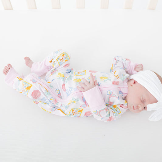 A sleeping newborn baby in a white crib, wearing a light pink and white long-sleeve footed sleeper with a colorful pastel summer beach travel pattern and a matching white bow headband.