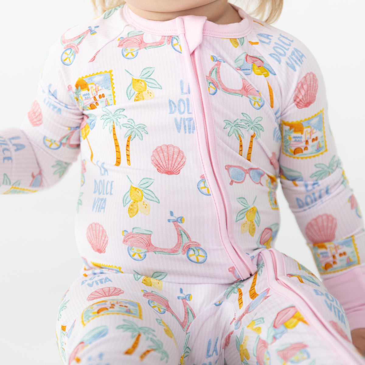 A close-up of a baby wearing a long-sleeve pink zip-up sleeper with a colorful 'La Dolce Vita' tropical print featuring palm trees, lemons, and scooters.