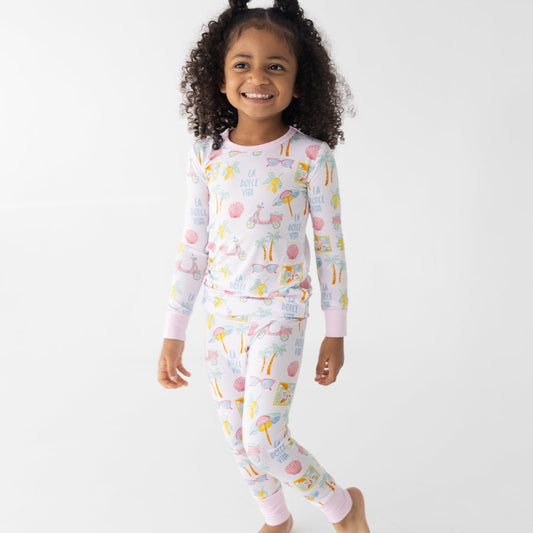 A young girl with curly dark hair wearing a light pink long-sleeve pajama set with a fun bright summer beach travel pattern, standing barefoot against a plain white background.