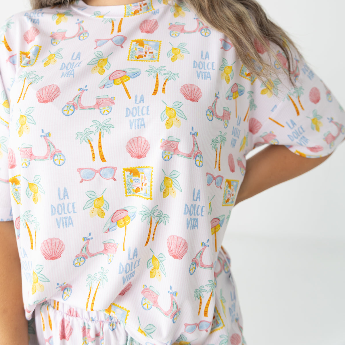 A close-up of a woman wearing a white short-sleeve pajama top with a colorful tropical beach-themed print featuring palm trees, seashells, sunglasses, and the text 'LA DOLCE VITA'.