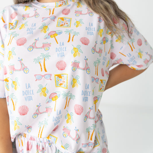 A close-up of a woman wearing a white short-sleeve pajama top with a colorful tropical beach-themed print featuring palm trees, seashells, sunglasses, and the text 'LA DOLCE VITA'.