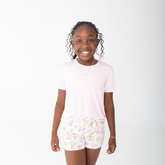 A young girl smiling and wearing a light pink short-sleeve t-shirt and white shorts with a colorful travel beach print, set against a plain white studio background.