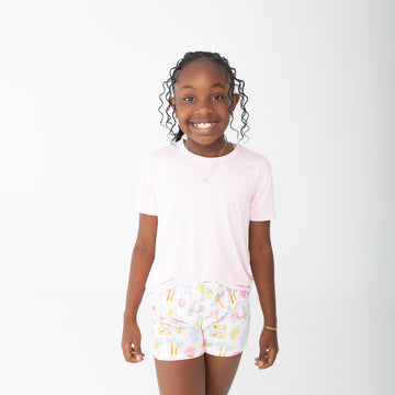 A young girl smiling and wearing a light pink short-sleeve t-shirt and white shorts with a colorful travel beach print, set against a plain white studio background.