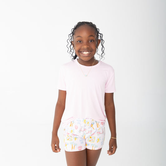 A young girl smiling and wearing a light pink short-sleeve t-shirt and white shorts with a colorful travel beach print, set against a plain white studio background.