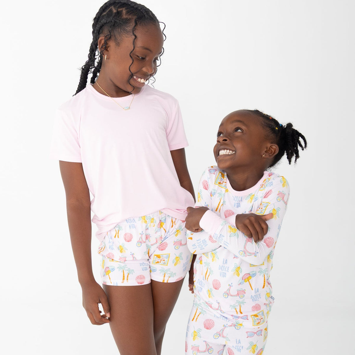 Two young girls modeling matching pajama sets against a plain white studio background. The older girl wears a light pink short-sleeve T-shirt and white shorts with a colorful beach travel print, while the younger girl wears a matching long-sleeve pajama set in white with the same print and pink cuffs.