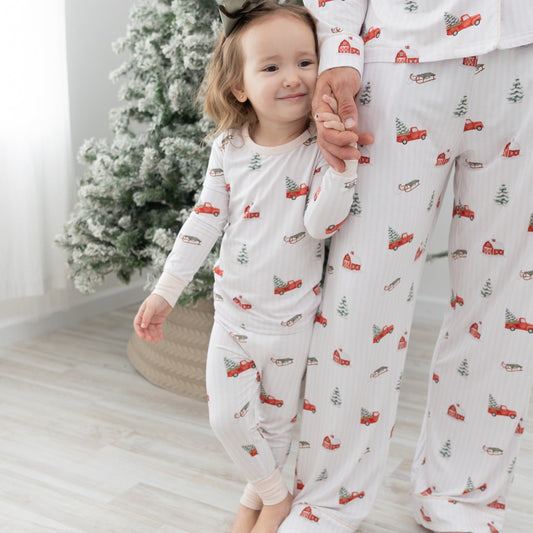 A toddler and an adult wearing matching white long-sleeve pajamas featuring a farmhouse, flocked christmas tree, green sleigh, red truck with a flocked christmas tree in the bed of the truck repeating pattern. standing barefoot in a bright room next to a decorated Christmas tree.