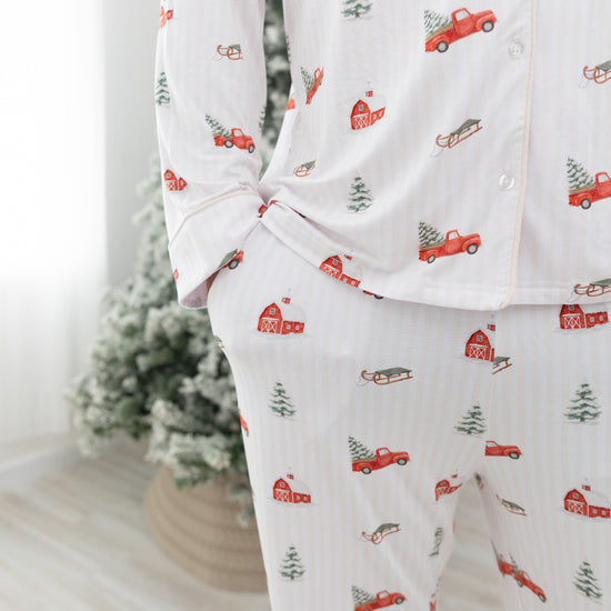 A person wearing a matching long-sleeve pajama top and pants set with a white base, subtle vertical stripe texture, and a repeating holiday print of red pickup trucks, red barns, evergreen trees, and sleighs. The background shows a softly blurred indoor setting with a Christmas tree.