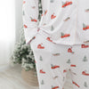 A person wearing a matching long-sleeve pajama top and pants set with a white base, subtle vertical stripe texture, and a repeating holiday print of red pickup trucks, red barns, evergreen trees, and sleighs. The background shows a softly blurred indoor setting with a Christmas tree.