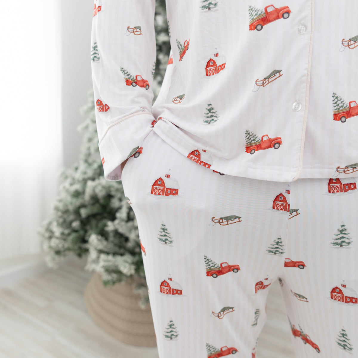 A person wearing a matching long-sleeve pajama top and pants set with a white base, subtle vertical stripe texture, and a repeating holiday print of red pickup trucks, red barns, evergreen trees, and sleighs. The background shows a softly blurred indoor setting with a Christmas tree.