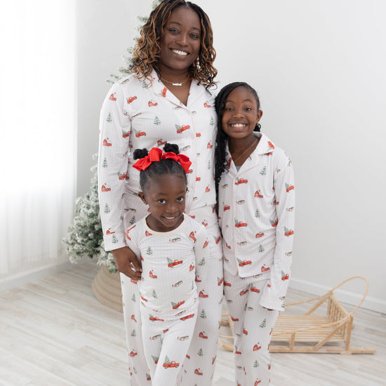 Three people wearing a long-sleeve white pajama set with a festive Christmas print featuring red pickup trucks, red barns, pine trees, and sleds, standing barefoot in a bright indoor room with light wood floors.