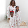 Three people wearing a long-sleeve white pajama set with a festive Christmas print featuring red pickup trucks, red barns, pine trees, and sleds, standing barefoot in a bright indoor room with light wood floors.
