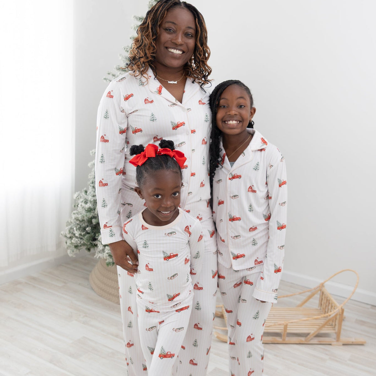 Three people wearing a long-sleeve white pajama set with a festive Christmas print featuring red pickup trucks, red barns, pine trees, and sleds, standing barefoot in a bright indoor room with light wood floors.