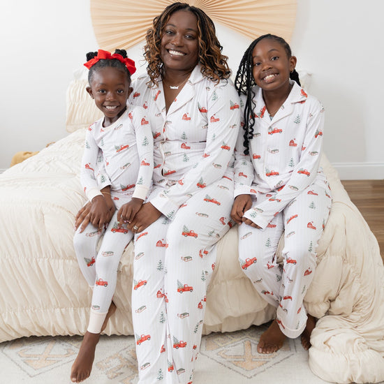 A woman and two children wearing matching long-sleeve white pajama sets with a festive Christmas print featuring red pickup trucks, red barns, pine trees, and sleds sitting together on a bed with beige bedding in a bright bedroom.