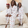 A woman and two children wearing matching long-sleeve white pajama sets with a festive Christmas print featuring red pickup trucks, red barns, pine trees, and sleds sitting together on a bed with beige bedding in a bright bedroom.