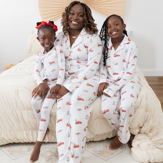 A woman and two children wearing matching long-sleeve white pajama sets with a festive Christmas print featuring red pickup trucks, red barns, pine trees, and sleds sitting together on a bed with beige bedding in a bright bedroom.