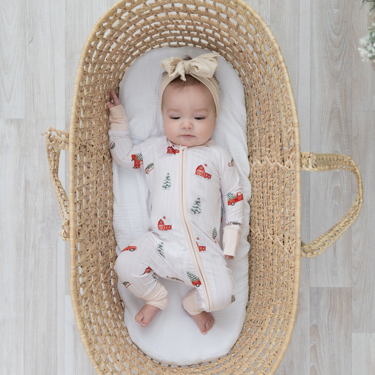 A baby wearing a white base with cream colored accent romper featuring a farmhouse, flocked christmas tree, red truck with a flocked christmas tree in the bed of the truck repeating pattern. Baby is lying inside a woven Moses basket on a light wood floor.