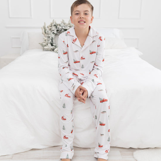 A young boy wearing a white long-sleeve pajama set with a red truck and green pine tree pattern, sitting on a white bed in a bright bedroom.