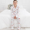 A young boy wearing a white long-sleeve pajama set with a red truck and green pine tree pattern, sitting on a white bed in a bright bedroom.