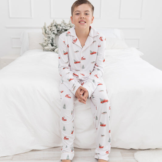 A young boy wearing a white long-sleeve pajama set with a red truck and green pine tree pattern, sitting on a white bed in a bright bedroom.