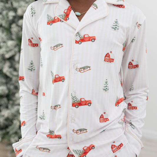 A person wearing white long-sleeve pajamas with a festive Christmas print featuring red pickup trucks, red barns, pine trees, and sleds. The image is cropped from the neck down, showing the pajama top and matching bottoms.