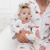 A baby wearing a matching white pajama set featuring a farmhouse, flocked christmas tree, green sleigh, red truck with a flocked christmas tree in the bed of the truck repeating pattern. Sitting on a man's lap in the same printed long sleave button pajamas.