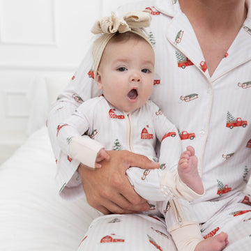 A baby wearing a matching white pajama set featuring a farmhouse, flocked christmas tree, green sleigh, red truck with a flocked christmas tree in the bed of the truck repeating pattern. Sitting on a man's lap in the same printed long sleave button pajamas.