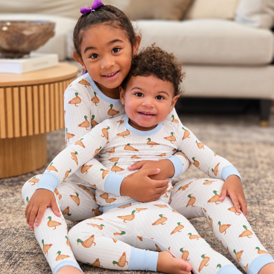  Photo shows a brother and sister sitting together in the living room on a rug. They're embracing and smiling at the camera. Both are wearing Monkeeroos Mallards toddler pjs 
