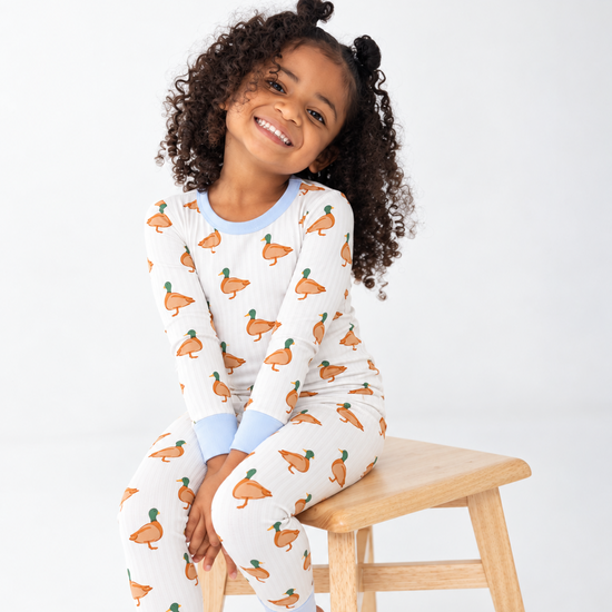 A little girl sitting on a stool smiling wearing Monkeeroos Mallards toddler pjs. It shows a cream striped background with blue cuffs with mallard ducks all over