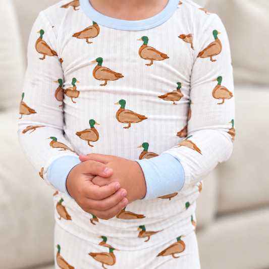 A toddler standing in front of a couch wearing Monkeeroos Mallards toddle pjs. It shows a cream striped background with blue cuffs with mallard ducks all over
