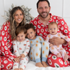 A family of four wearing matching Christmas-themed pajamas sits together on a white bed. The father and mother wear red pajamas with a white Santa Claus pattern, while the two children wear light-colored pajamas with gingerbread motifs.