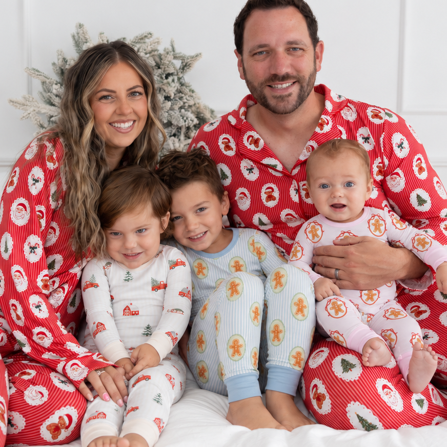 A family of four wearing matching Christmas-themed pajamas sits together on a white bed. The father and mother wear red pajamas with a white Santa Claus pattern, while the two children wear light-colored pajamas with gingerbread motifs.